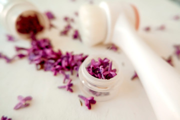 White jar with lilac petals on a white background, facial massager, alternative medicine and skin care.