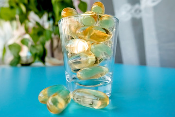 A glass cup with omega 3 vitamins stands on a blue table near the window, near a birch tree,.