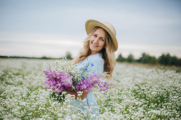 beautiful blonde girl in a field of daisies. woman in a blue dress in a field of white flowers. girl with a bouquet of daisies. summer tender photo in the village. wildflowers. girl in a straw hat