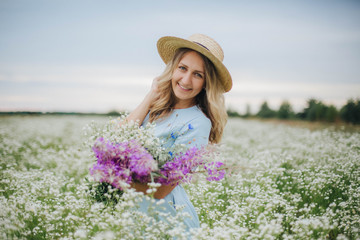 beautiful blonde girl in a field of daisies. woman in a blue dress in a field of white flowers. girl with a bouquet of daisies. summer tender photo in the village. wildflowers. girl in a straw hat