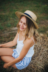 beautiful girl in a straw hat on a field with straw bales. girl sitting on the hay. blonde in the summer in a field with a hat. life in the countryside, outdoors. eco project. clean environment