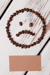 Sad smiley face made of coffee beans. Beige paper for copyspace on white wooden planks.
