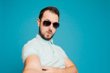 A young bearded man is fooling around at the camera on a blue background. The guy takes a selfie and builds funny faces