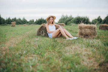 Naklejka na ściany i meble beautiful girl in a straw hat on a field with straw bales. girl sitting on the hay. blonde in the summer in a field with a hat. life in the countryside, outdoors. eco project. clean environment