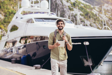 Young man walking by the harbor of a touristic sea resort with boats on background, holding digital tablet.