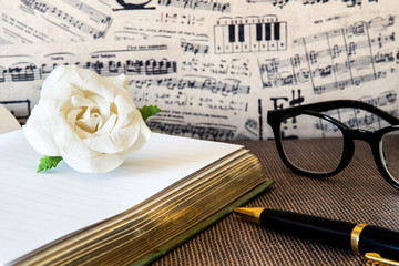 Fototapeta na wymiar White paper rose on note book with black and gold pen against musical note wallpaper.