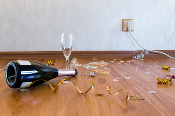 A bottle of champagne and empty glasses left after party on the floor with confetti and serpentine.