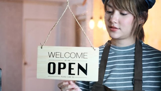 Business owner attractive young Asian woman in apron hanging we're open sign on front door smiling welcoming clients to new cafe. Beautiful Young Cafe Owner Turning Storefront Sign From Close to Open.