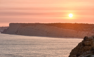 Beautiful sunset over the cliffs of the beach, city of tourist apartments and wind mills. Concept of tourism and travel. Algarve, Portugal