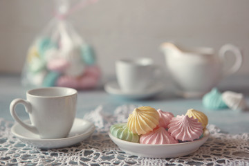 Multi-colored meringues in pastel shades with cups and a jug on a light textured wooden background. The concept of Breakfast with tea or coffee, home comfort and tenderness. Close-up, selective focus