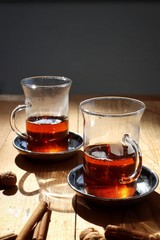 Two glass cups of tea on the wooden table out on the balcony on a sunny day. Some orange peel, cinnamon and biscuits the side. Vertical, closeup.