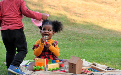 Adorable African girl enjoys to playing her toys in park or garden during a picnic with her family.