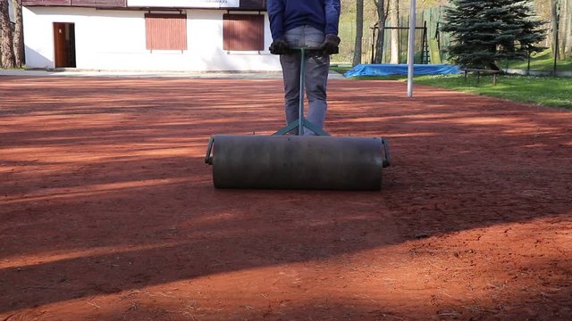 Young summer worker prepared clay court for tournament and league volleyball matches. Labourer pulls a heavy roller behind him and suffocates the clay. Gardening. Adjust the surface of the court
