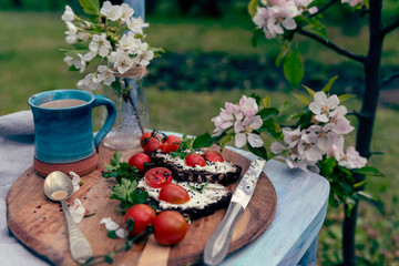 Healthy breakfast on rural chair in the garden. Vegetarian food with mug of coffeee and blooming flowers outdoors.  Homemade slice of bread with red tomatoes and cheese with spoon and knife. Close up.