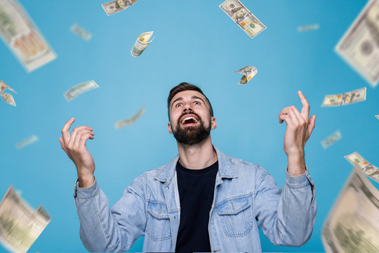 Young cheerful man in black t-shirt and denim jacket. Guy with beard expresses happiness. Blue background. Money rain. Falling hundred dollar banknotes.