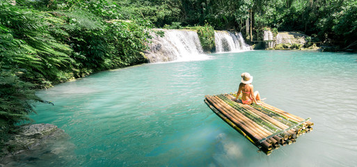 Woman in bikini and hat sitting on bamboo raft and enjoying view on waterfall. Travel and vacation...
