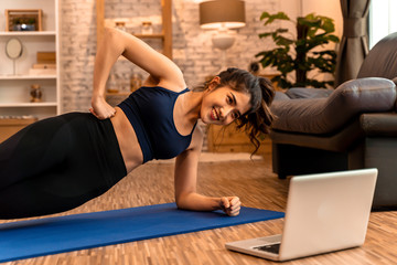 20s young Asian woman in sportswear doing stretching exercises while watching fitness training class on computer laptop online. Healthy girl exercising in living room with sofa couch in background.