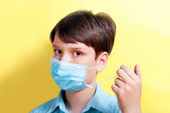 A boy in a blue shirt on a yellow background puts on a blue medical mask. Concept protection against viruses, bacteria, flu, colds, infections, COVID-19. - Powered by Adobe