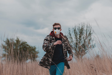 A man in a protective mask and camouflage jacket runs across the field in nature. Jogging during self-isolation during the coronavirus epidemic.