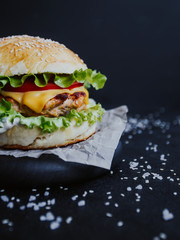 Appetizing fresh homemade burger, with chicken cutlet, lettuce, tomatoes, cheese and sauce. On a wooden board on a black background