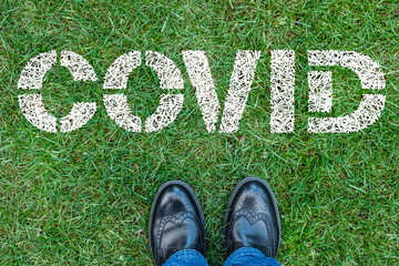 Black shoes standing next to covid sign. Covid sign painted on on green grass.