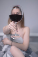 happy beautiful slender half-naked girl shows us a huge glass of red wine, in bed with gray sheets in triangles - vertical photo
