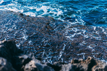 A piece of the Mediterranean Sea with foam and stones of dark blue color, with blurry stones in the foreground. Nice seascape