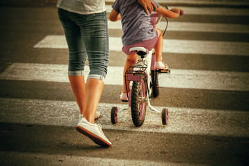Woman with son crossing the road in the city. Mother goes pedestrian crossing with childr on bicycle. Back view.