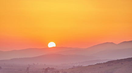 Panorama of beautiful mountains covered with trees at dawn in summer. Bright large orange sun disc. Solar reflection. Flash of the sun. Lens flare. Nature, leisure for the tourist.