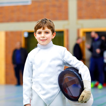 Little Kid Boy Fencing On A Fence Competition. Child In White Fencer Uniform With Mask And Sabre. Active Kid Training With Teacher And Children. Healthy Sports And Leisure.