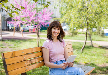 Obraz premium A young girl holds a tablet in her hands, headphones in her ears. Cute girl listening to music through headphones on a tablet. A girl in a pink t-shirt and jeans sits on a bench on the street.