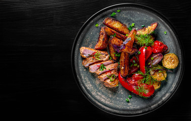 fried meat with potatoes, pepper, tomatoes, herbs and spices in plate on black wooden table background