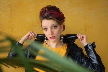 Glamorous lady in punk rock style party clothes with dark pink hair in casual dress, black leather jacket, and long earrings on yellow background