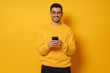 Young handsome man holding smartphone in hands, looking at camera with positive smile, enjoying communication with friends, isolated on yellow background