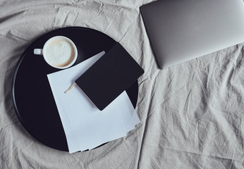 Laptop laying on blanket, next to notebook, sheets of paper and cup of cappuccino on black round tray, top view