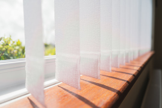 White vertical window blinds slats with cordless glued weighted pockets on the end casting shadows on the wooden window sill.