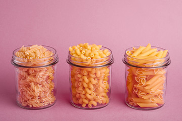 Three glass jars with variety of uncooked golden wheat pasta on minimal pink background, angle view