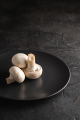 Three champignon mushrooms healthy food on black plate on dark black textured background, angle view copy space