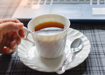 A hand and a white coffee cup handle in sunshine morning  on the desk at home, Notebook computer and work equipment, Normal practice by working from home during covid-19 outbreak around the world.