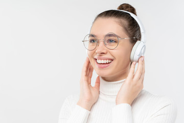 Teen girl listening to favorite music tracks, touching wireless headphones she is wearing, looking aside and smiling happily, isolated on gray background