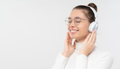 Horizontal banner of young smiling female in glasses, enjoying music through wireless headphones, eyes closed with pleasure, isolated on gray background with copy space