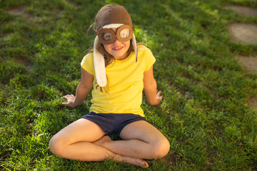 Authentic shot of cute happy little girl with aviator hat is sitting on a green grass and smiling in a sunny day. Concept:adventure,freedom, dreams, imagination, childhood,happiness, authenticity