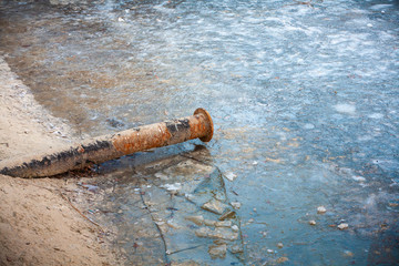 Cloudy winter day. The lake is covered with ice. On the sandy shore is an old rusty pipe.
