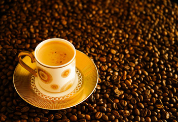 White cup of coffee, foamy espresso on gold saucer on roasted brown coffee beans background.