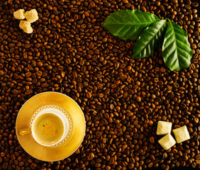 Naklejka premium Roasted coffee beans background white cup on golden saucer with foamy espresso, top view. On beans fresh green coffee leaves, brown sugar cubes, slices of chocolate.
