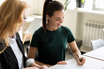 Happy young attractive businesswoman writing signing paper document with mature female partner....