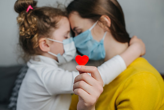 Mother And Little Daughter Embrace Bonding, Wearing Facial Medical Mask, Holding Red Heart A Way To Show Appreciation And To Thank All Essential Employees During Covid-19 Pandemic. Selective Focus