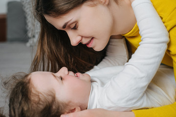 Obraz na płótnie Canvas Side view of affectionate mother cuddling little adorable daughter, enjoying sweet tender moment in bedroom. Smiling woman looking at eyes of adorable small child girl, laughing having fun at home