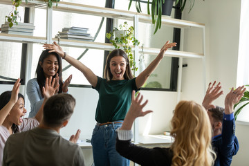 Happy attractive businesswoman conduct training with diverse colleagues. Diverse team celebrating business success at meeting. Smiling female mentor with raised hands congratulating colleagues.