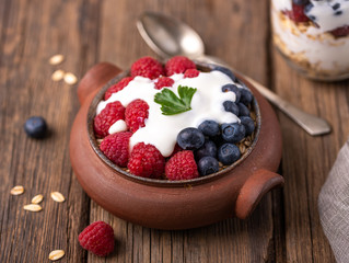 Granola with white yogurt with raspberries and blueberries in ceramic bowl on natural wooden desk.